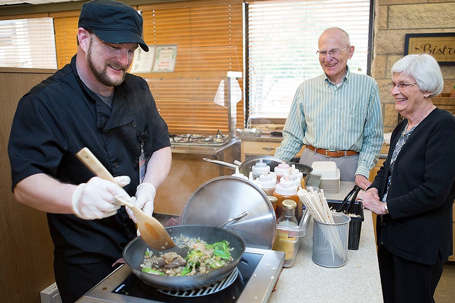 Male Chef Cooking In Pan With Two Residents Watching Male Chef Cooking In Pan With Two Residents Watching